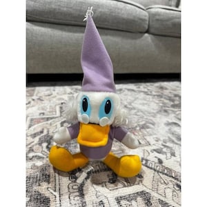 May include: A plush toy featuring a character with a white face, blue eyes, and a yellow beak. The character wears a purple hat and a purple body with yellow feet. The toy is sitting on a patterned rug.