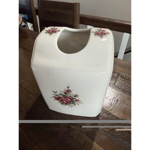 May include: A white ceramic tissue box cover with a rectangular shape and rounded top edges. The surface features a floral pattern with clusters of red roses and green leaves centered on the front and on both sides of the top opening.