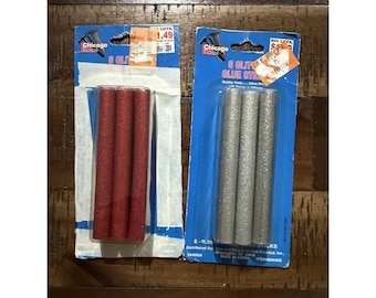 Pode incluir: Duas embalagens de bastões de cola com glitter Chicago Tool 6 Glitter. Uma embalagem contém bastões de cola com glitter vermelhos e a outra bastões de cola com glitter prateados. As embalagens estão sobre uma superfície de madeira.