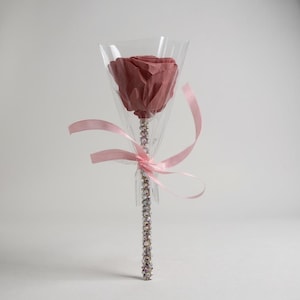 Puede incluir: Una rosa preservada en un cono de plástico transparente, atada con una cinta rosa. El tallo está adornado con brillantes pedrería. La rosa es de color malva apagado y el fondo es blanco.