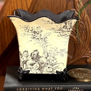 May include: A decorative vase with a black scalloped rim and a cream-coloured body featuring a black and white pastoral scene. The vase sits atop a stack of books with titles like "Forgiving What You Can't Forget" and "Mysteries of Mankind".