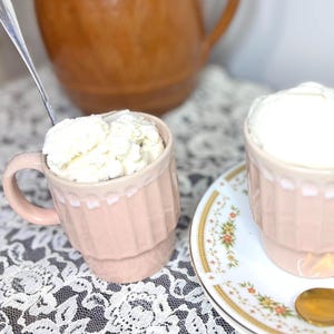 Könnte beinhalten: Zwei rosa Tassen gefüllt mit Eiscreme, eine mit einem Löffel. Die Tassen stehen auf einer weißen Spitzendecke, mit einer Blumenschale und einem goldenen Löffel. Eine braune Kanne mit rosa Blumen ist im Hintergrund.
