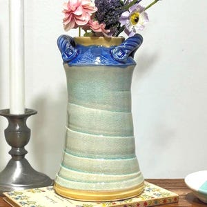 May include: A handmade ceramic vase with a blue rim and handles, filled with pink, purple, and white flowers. The vase sits on a wooden tray with a candle and a small book. Two small blue and white coasters are also on the tray.