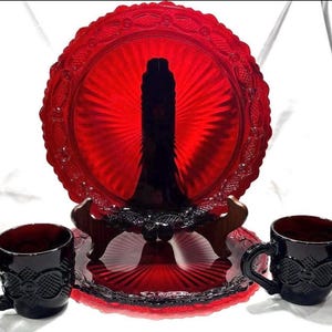 May include: A red glass serving plate with a black center, displayed on a decorative stand, flanked by two black mugs. The plate has a scalloped edge and a radial design. The mugs have ornate handles and detailing.