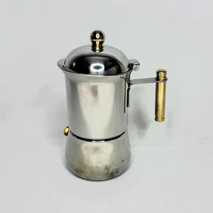 Könnte beinhalten: Ein Espressokocher aus Edelstahl mit goldfarbenem Griff und Deckelknopf. Der Kaffeezubereiter hat ein klassisches Design mit rundem Boden und einem Deckel, der sich öffnen lässt. Der Griff ist an der Seite des Topfes befestigt.