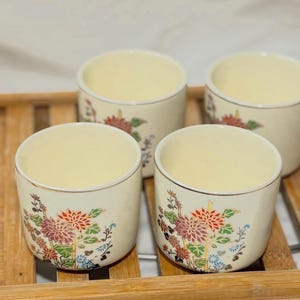 May include: Four cream-colored ceramic teacups with floral designs in red, pink, and green. The cups have a gold rim and are arranged on a wooden tray. The floral pattern includes chrysanthemums.