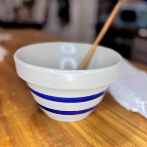 Puede incluir: Un cuenco de cerámica blanca con dos rayas azules horizontales. El cuenco tiene un borde ancho y está sobre una superficie de madera. Una cuchara de madera descansa dentro del cuenco. El cuenco se utiliza probablemente para hornear o cocinar.