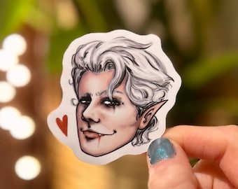Könnte beinhalten: Ein Aufkleber mit einer stilisierten Illustration einer Person mit weißen Haaren, spitzen Ohren und Fangzähnen. Die Person hat dunkles Augen-Make-up und ein rotes Herz. Der Aufkleber wird von einer Hand mit einem blauen Nagel gehalten.