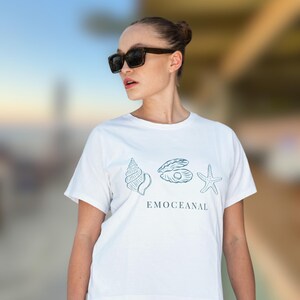 Puede incluir: Camiseta blanca con ilustraciones turquesas de una concha, ostra y estrella de mar, con la palabra "EMOCEANAL" impresa debajo. La camiseta es de manga corta.