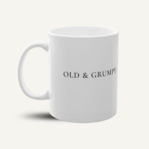 Könnte beinhalten: Eine weiße Keramik-Tasse mit schwarzem Henkel und dem Text "But First, Coffee" in fetter, schwarzer Schrift. Die Tasse steht vor einem neutralen Hintergrund, der ihr schlichtes Design hervorhebt.