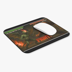 May include: Black rectangular mousepad featuring a fantasy battle scene. The image depicts a green creature labeled "The Van Meter Visitor" facing off against "Bigfoot." The word "FIGHT!" is in red. A white computer mouse rests on the pad.