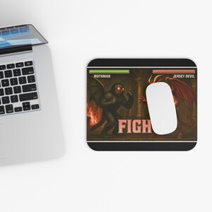 May include: Black rectangular mousepad featuring a graphic of Mothman versus Jersey Devil. The word "FIGHT" is in the center. A white computer mouse is on the right side of the mousepad.