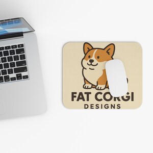 May include: A beige mousepad with a cartoon corgi illustration in brown and white, and the text "FAT CORGI DESIGNS". A white computer mouse rests on the mousepad, next to a laptop.