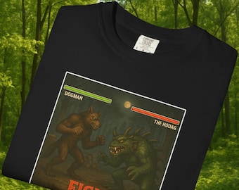 May include: Black t-shirt featuring a graphic of a Dogman versus The Hodag in a forest setting. The design includes the word "FIGHT!" and health bars for each creature. The shirt is made by Comfort Colors.