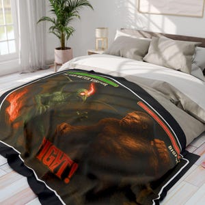 Puede incluir: Una manta negra con una escena de batalla de fantasía. Representa una criatura alada verde y un Bigfoot marrón. La manta tiene el texto rojo "FIGHT!" y el texto blanco "THE VAN METER VISITOR" y "BIGFOOT".