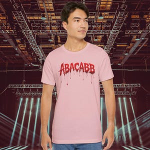 May include: A light pink t-shirt with the word "ABACABB" in red, dripping letters. The shirt is a short-sleeved crew neck style. The background is a concert stage with lights and metal structures.