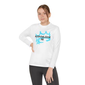 May include: White long-sleeve shirt with the text "ACKNOWLEDGE ME" in a blue, graffiti-style design. The shirt is paired with black leggings. The overall style is casual and modern.