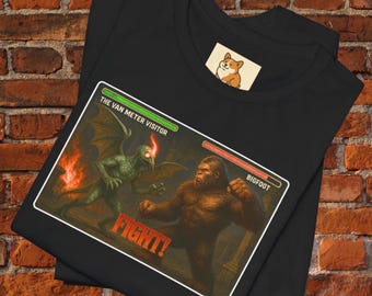 May include: Black t-shirt featuring a graphic of a battle scene between a winged creature and Bigfoot, with the text "FIGHT!". The graphic includes the text "The Van Meter Visitor" and "Bigfoot". A small corgi logo is on the shirt.