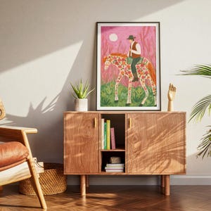Op de afbeelding: Een ingelijste kunstdruk toont een persoon op een paard versierd met bloemmotieven. Het kunstwerk heeft een roze en groene achtergrond met een volle maan. De ruiter draagt een hoed en een vest. De print wordt getoond op een houten kast met gouden handgrepen.