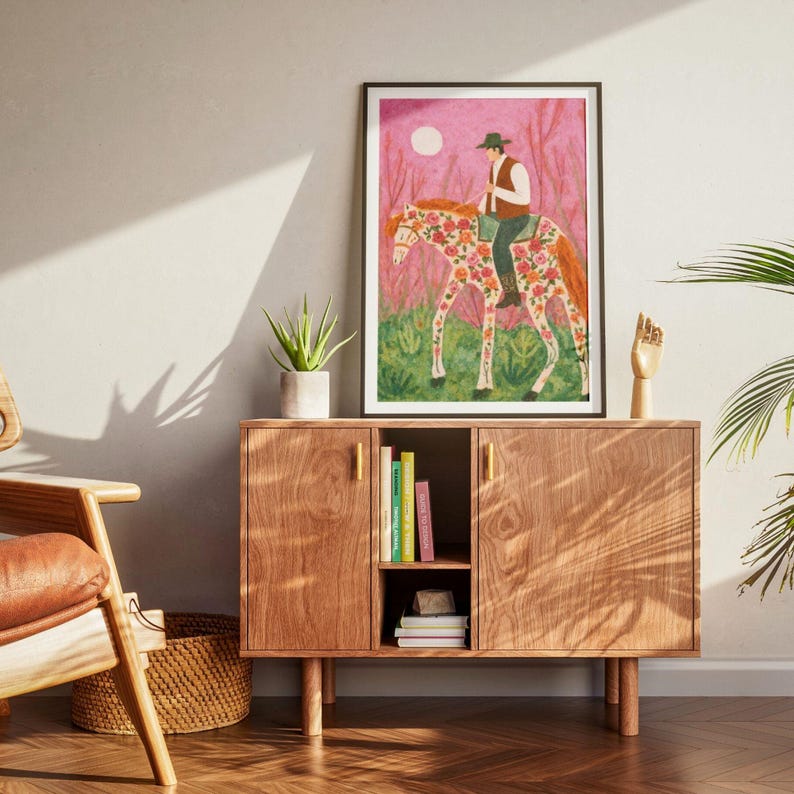 Op de afbeelding: Een ingelijste kunstdruk toont een persoon op een paard versierd met bloemmotieven. Het kunstwerk heeft een roze en groene achtergrond met een volle maan. De ruiter draagt een hoed en een vest. De print wordt getoond op een houten kast met gouden handgrepen.