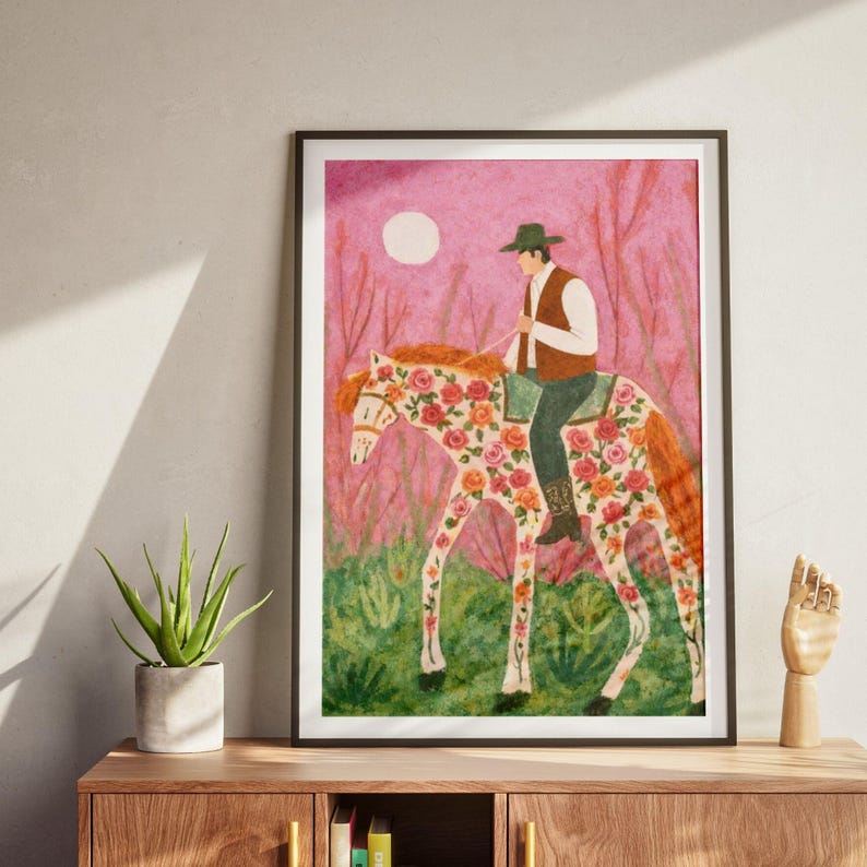 Op de afbeelding: Een ingelijste kunstdruk toont een persoon op een paard met bloemmotieven. Het kunstwerk heeft een roze achtergrond met een volle maan en gestileerde bomen. De ruiter draagt een hoed en vest. De vacht van het paard is versierd met rode en roze rozen.