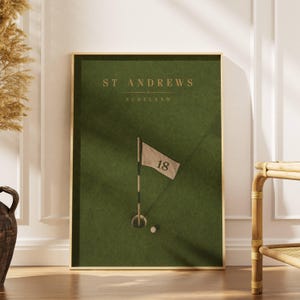 May include: A framed vertical wall art print featuring a dark green background with the text ST ANDREWS SCOTLAND in gold lettering. The design includes a golf hole with a beige flag marked with the number 18 and a white golf ball on the green.