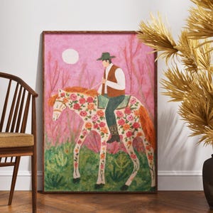 Op de afbeelding: Een ingelijste kunstdruk toont een persoon op een paard versierd met bloemen. De ruiter draagt een groene hoed en vest. De achtergrond is roze met een witte maan en hoge planten. Het paard heeft een witte basis met oranje en roze bloemmotieven.