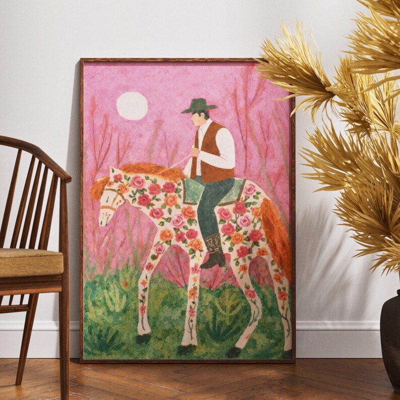 Op de afbeelding: Een ingelijste kunstdruk toont een persoon op een paard versierd met bloemen. De ruiter draagt een groene hoed en vest. De achtergrond is roze met een witte maan en hoge planten. Het paard heeft een witte basis met oranje en roze bloemmotieven.