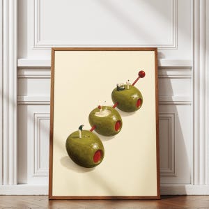May include: A framed art print featuring three green olives skewered on a red cocktail pick. Tiny figures of golfers are positioned on the surface of each olive, with one golfer near a golf cart, creating a surreal scene on a cream background.