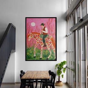 Op de afbeelding: Een ingelijst schilderij toont een persoon te paard versierd met bloemmotieven. De achtergrond is roze met een witte maan. De ruiter draagt een hoed en een vest. Het kunstwerk wordt tentoongesteld in een kamer met een tafel en stoelen.