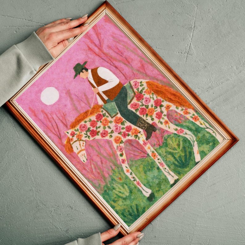 Op de afbeelding: Een ingelijste kunstdruk met een ruiter op een paard versierd met bloemmotieven. Het kunstwerk heeft een roze achtergrond met een witte maan en groen gebladerte. De ruiter draagt een hoed, vest en donkere broek. De lijst is bruin.