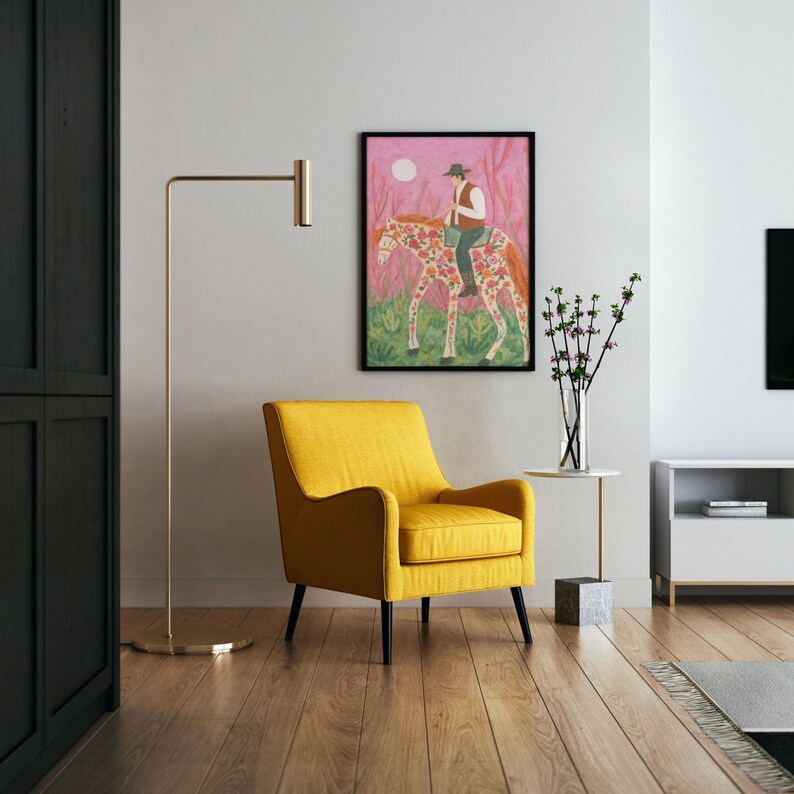 Op de afbeelding: Een ingelijste kunstdruk toont een persoon op een paard met bloemmotieven. Het kunstwerk heeft een roze achtergrond, een witte maan en groen gebladerte. Voor het kunstwerk staat een gele fauteuil met zwarte poten.