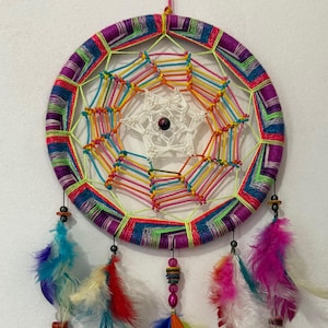 May include: A colorful dreamcatcher with a circular woven frame in purple, blue, and green, with a rainbow-colored web in the center. It features hanging feathers in shades of blue, orange, pink, and white, and small beads.