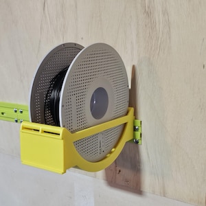 Peut inclure: Un support jaune pour bobine de filament d'imprimante 3D monté sur une surface en bois. Le support a une conception incurvée et contient deux bobines, une noire et une grise. Le support est fixé au mur avec des vis.