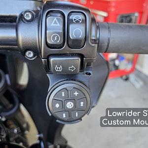 May include: Close-up of a motorcycle handlebar with black controls and a custom mount. The mount holds a circular control panel with buttons. The text "Lowrider ST Custom Mount" is visible.