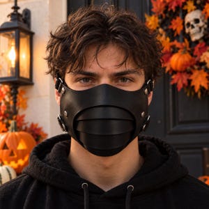 Peut inclure: Un masque facial noir présentant un design segmenté et superposé en matériau lisse. Le masque est fixé par des rivets métalliques et des anneaux latéraux, couvrant la zone du nez et de la bouche avec une configuration de panneaux structurés.