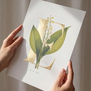 May include: A print featuring the letter "E" in gold foil, with a floral design of green leaves and white flowers. The date "06 / 03 / 2023" is printed at the bottom. The artwork is held by two hands.
