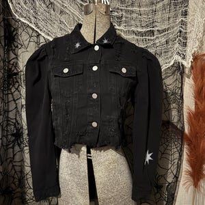 May include: A black denim jacket featuring a distressed finish, silver-toned buttons, and puff sleeves. The jacket is adorned with small, white, hand-painted star motifs on the collar points and on the lower portion of the left sleeve.