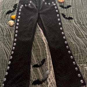 May include: Dark gray denim pants with a leopard print pattern and white skull accents along the side seams and pockets. The pants have a flared leg and are decorated with small white skulls. Halloween-themed apparel.