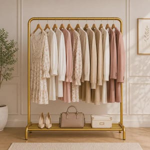 以下が含まれることがあります： ゴールドの金属製衣類ラックには、白、ベージュ、ダスティピンクの色合いのドレス、ブラウス、ジャケットが並んでいます。下の棚には、ベージュのパンプス、構造的なハンドバッグ、長方形の収納ボックスが置かれています。