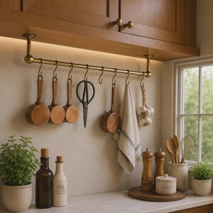 Può includere: Una barra da cucina in ottone montata sotto pensili in legno con ganci a S che sostengono tre misurini in rame, forbici nere, un piccolo pentolino in rame e un canovaccio beige a righe. Un mazzo d'aglio è appeso alla barra.