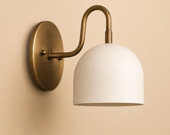 May include: A wall-mounted light fixture with a white, dome-shaped shade and a curved brass arm. The fixture is attached to a round brass base. The design is minimalist and modern, suitable for various interior styles.