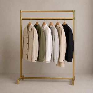 Könnte beinhalten: Ein goldfarbener Kleiderständer aus Metall mit zwei horizontalen Stangen, an dem sechs langärmelige Hemden auf Holzkleiderbügeln hängen. Die Hemden sind in einem Farbverlauf von Beige, Olivgrün, Weiß, Hellgrau und Tan bis Schwarz angeordnet.