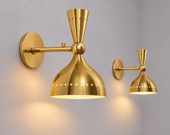 May include: Two gold-toned wall sconces featuring a dual-cone design. Each fixture has a circular wall mount, a horizontal arm, and a bell-shaped shade with a row of small circular perforations along the lower edge, emitting a warm light.