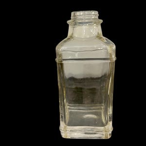 May include: A clear, rectangular glass bottle with a square base and a narrow neck. The bottle has a slightly curved design and is empty. The background is black.