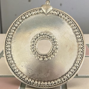 May include: Silver-toned round pendant with a heart-shaped bail. The pendant features a central circular design and a beaded border. The center has a small, circular mirror. The pendant is likely made of metal.