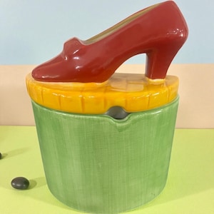 May include: A ceramic ashtray with a red high-heeled shoe on top. The ashtray is green with a yellow rim. The shoe is glossy red. The ashtray is a decorative item.