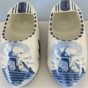 May include: Pair of white ceramic clogs with a blue windmill design. The clogs have a rounded toe and a hollow interior. The exterior is decorated with a blue and white windmill scene, with a blue border around the top edge.