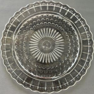 May include: Clear glass serving plate with a scalloped edge and a detailed, textured design. The plate features concentric circles and a central sunburst pattern, creating a decorative and elegant look. Ideal for serving appetizers or desserts.
