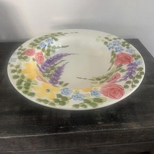 May include: A white ceramic serving bowl with a floral design. The rim is decorated with hand-painted flowers in shades of pink, yellow, blue, and purple, with green leaves. The bowl sits on a dark wooden surface.